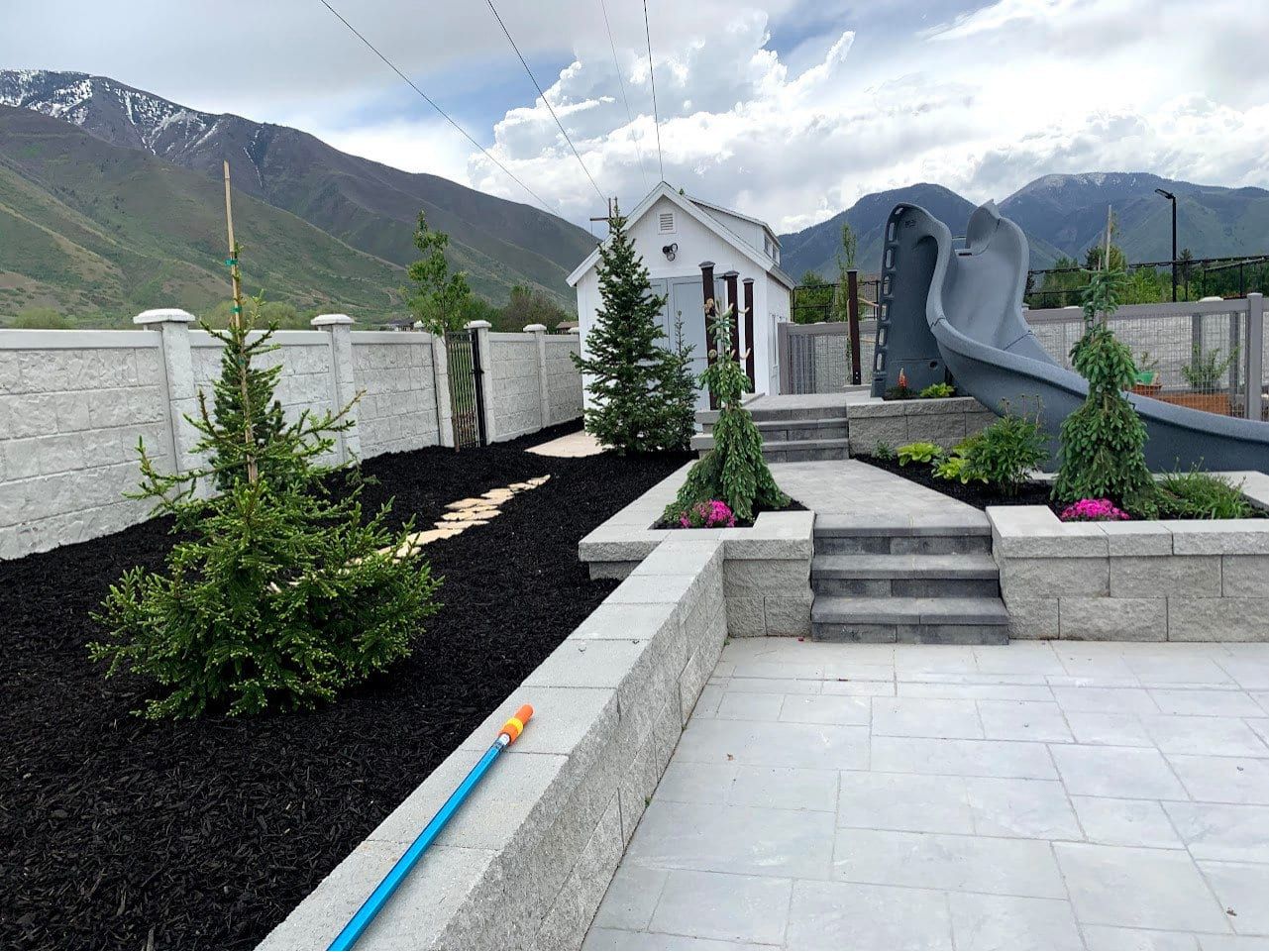 Block retaining wall and steps rising from a light gray patio, with young evergreens in black mulch and mountains behind